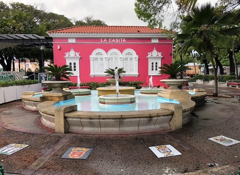 Fountain in Plaza Darsena surrounded by mosaic city crests embedded in the plaza.  Background is a bright pink building with decorative white accents