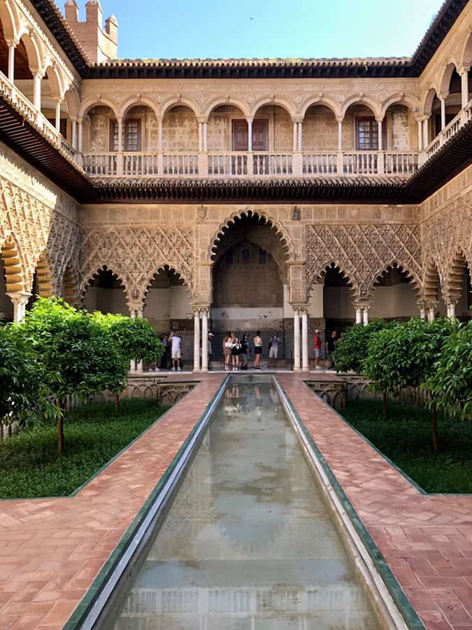 Intricate designs decorate the arches of the courtyard of the Royal Alcazar in Seville Spain.  A narrow pond runs down the center of the courtyard.